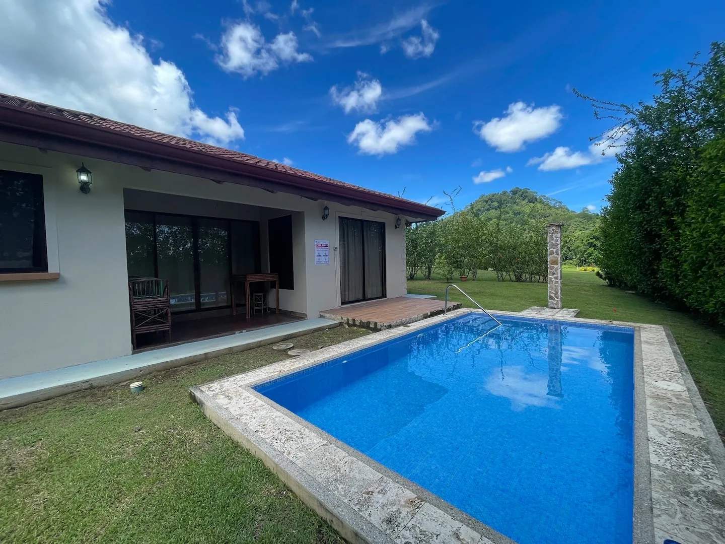 An outdoor swimming pool beside a low house, with a green lawn and trees.
