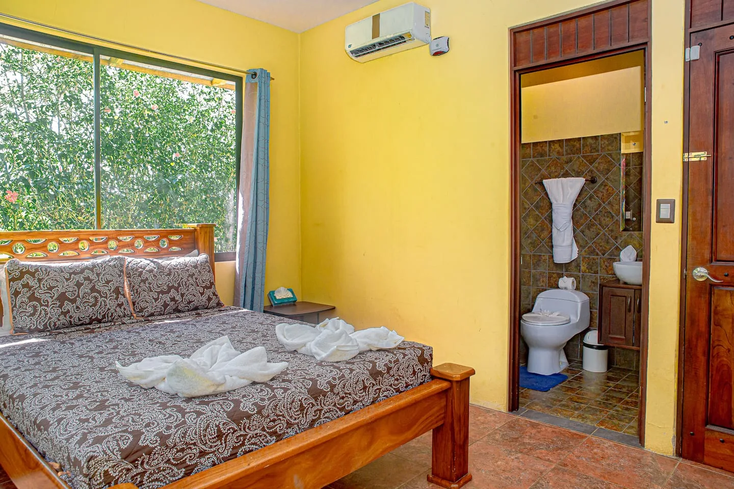 A bedroom with a wooden bed beside a window and an open bathroom doorway.