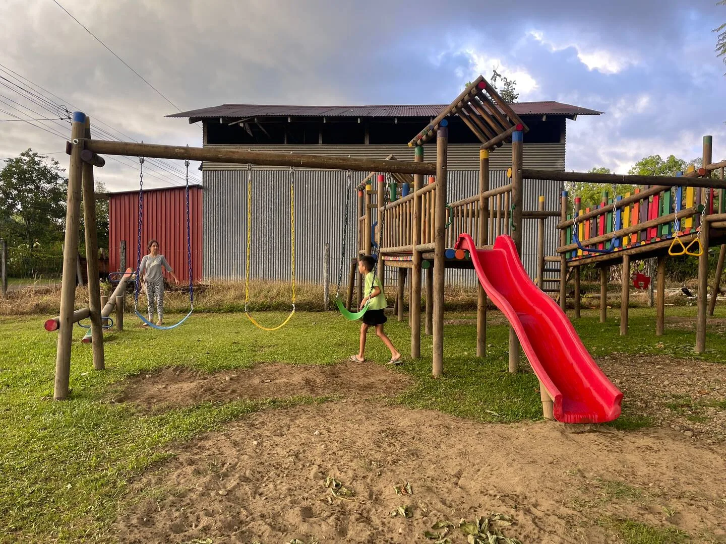 An outdoor playground has a wooden climbing structure, swings, and a bright red slide.