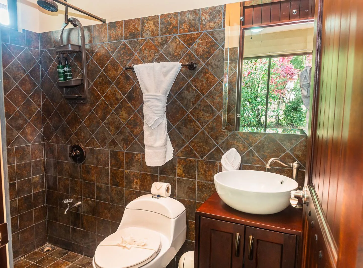 A tiled bathroom with a toilet, sink, and window overlooking greenery.