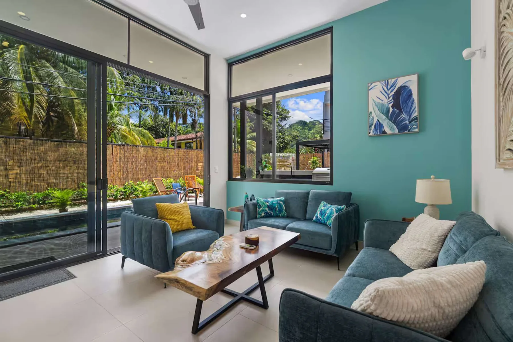 A living room with blue seating, a wooden coffee table, and large windows overlooking greenery.