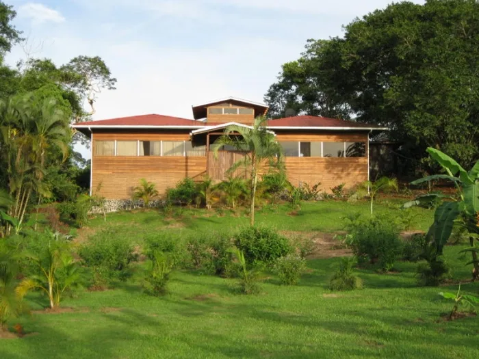 A low wooden house with a red roof sits among green lawns and trees.