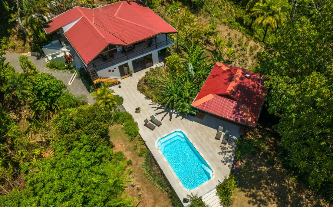 An aerial view of a house and a smaller building beside a swimming pool amid greenery.