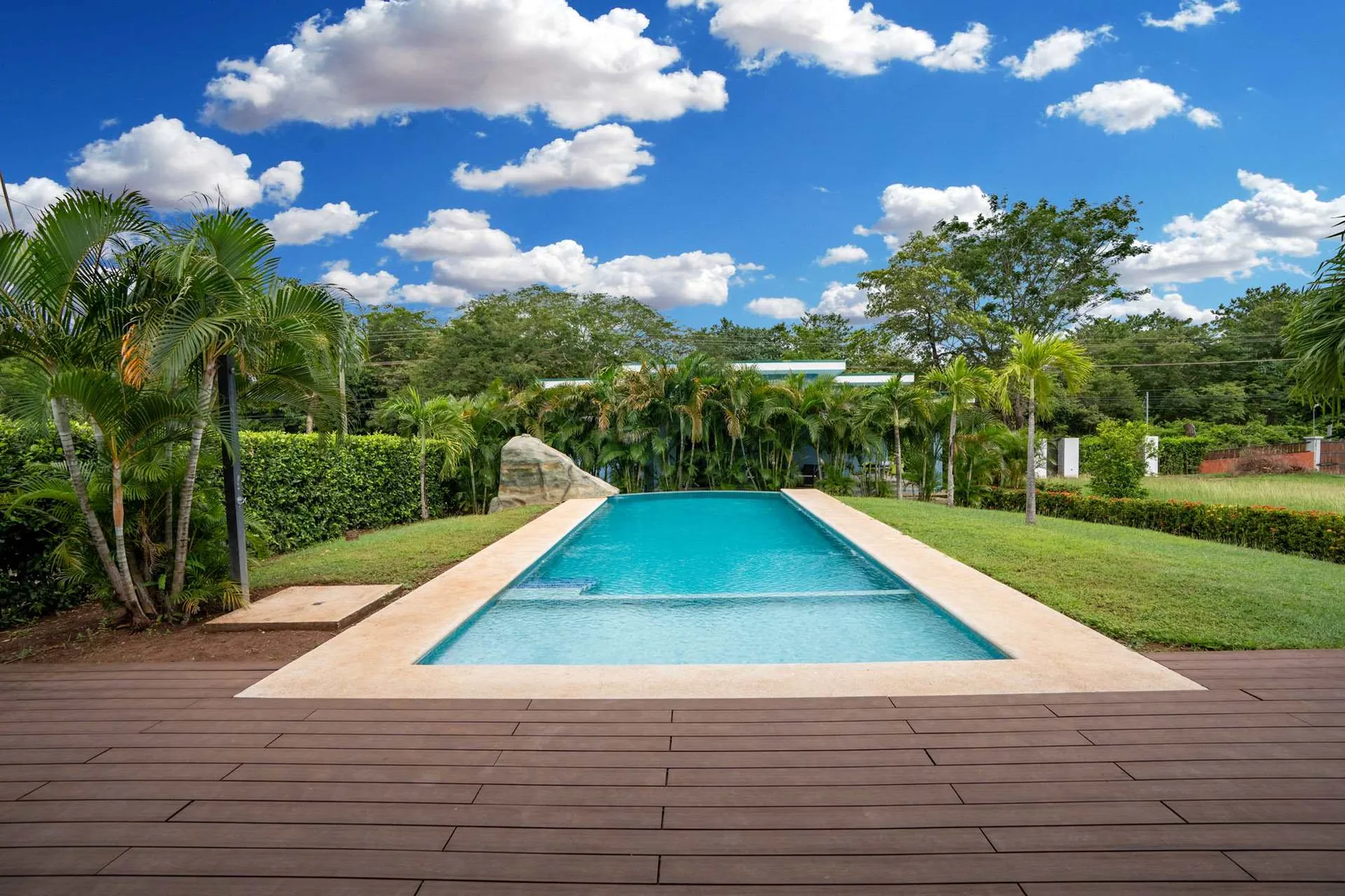 Outdoor swimming pool surrounded by a paved deck, lawn, and palm trees.