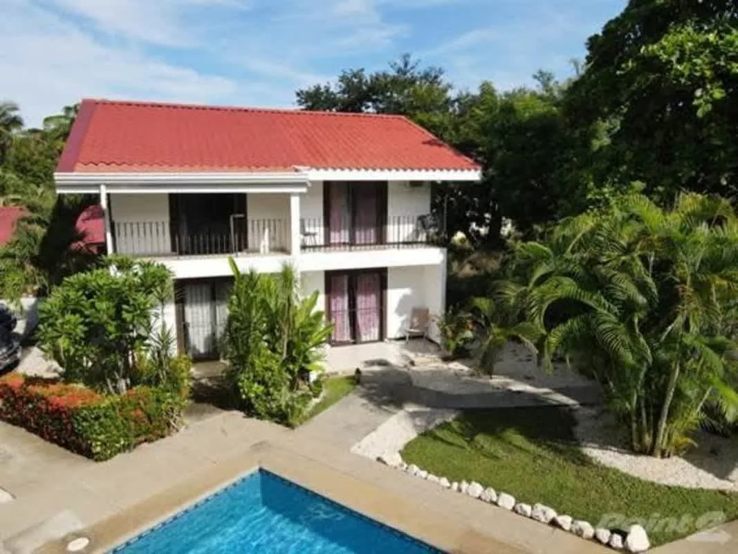 A two-story house with a red roof stands beside a swimming pool amid greenery.