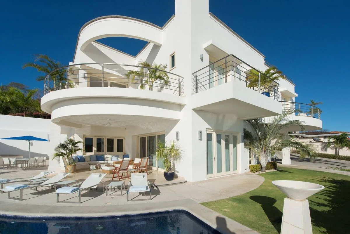 A modern white villa overlooks a swimming pool, with balconies and patio seating.