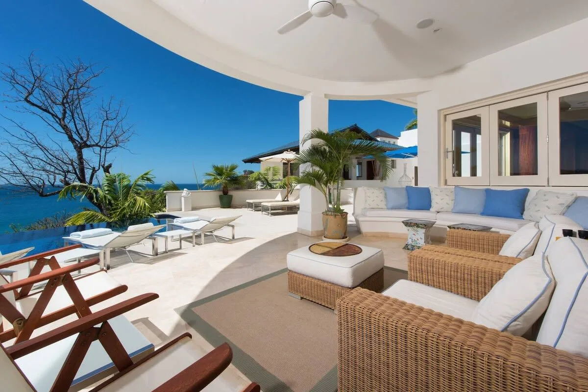 A seaside patio has cushioned seating, loungers, and a view of the water.