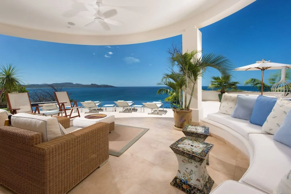 A covered seaside patio with cushioned seating, potted palms, and views of the ocean.