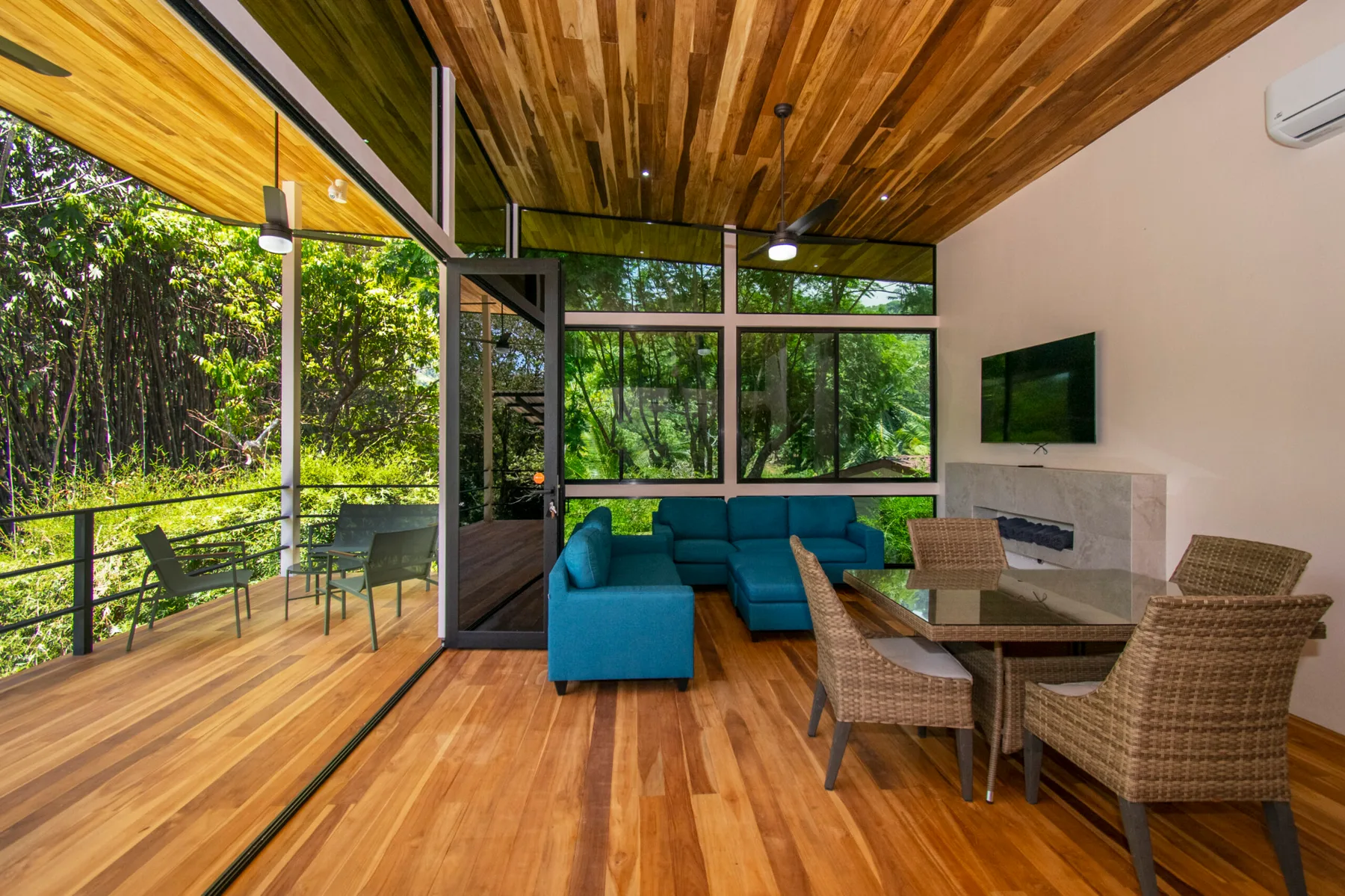 A living and dining area opens onto a leafy balcony, with seating and a television.