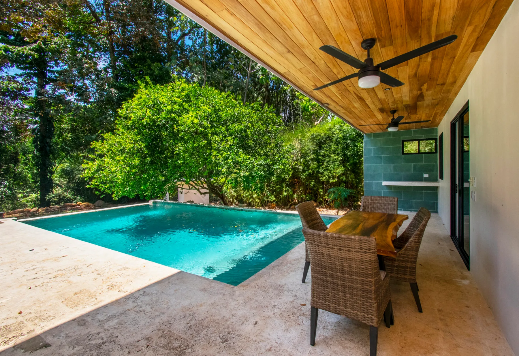 A covered patio with outdoor seating overlooks a swimming pool and leafy greenery.
