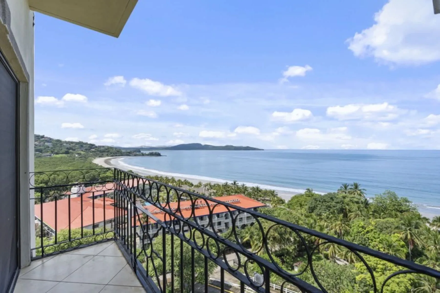 A balcony overlooks a curving shoreline and the ocean beneath a partly cloudy sky.
