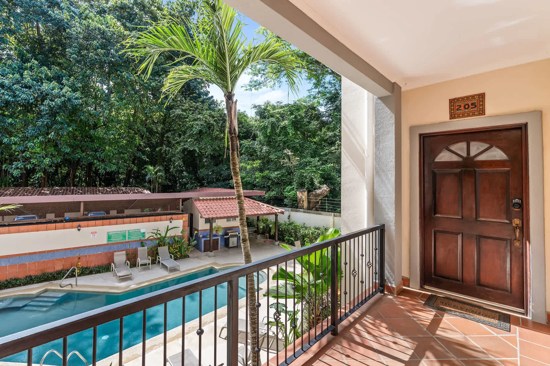 A balcony overlooks a swimming pool and lush greenery beside a wooden entrance door.