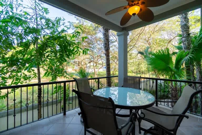 A covered outdoor seating area with a glass-top table overlooks leafy trees.