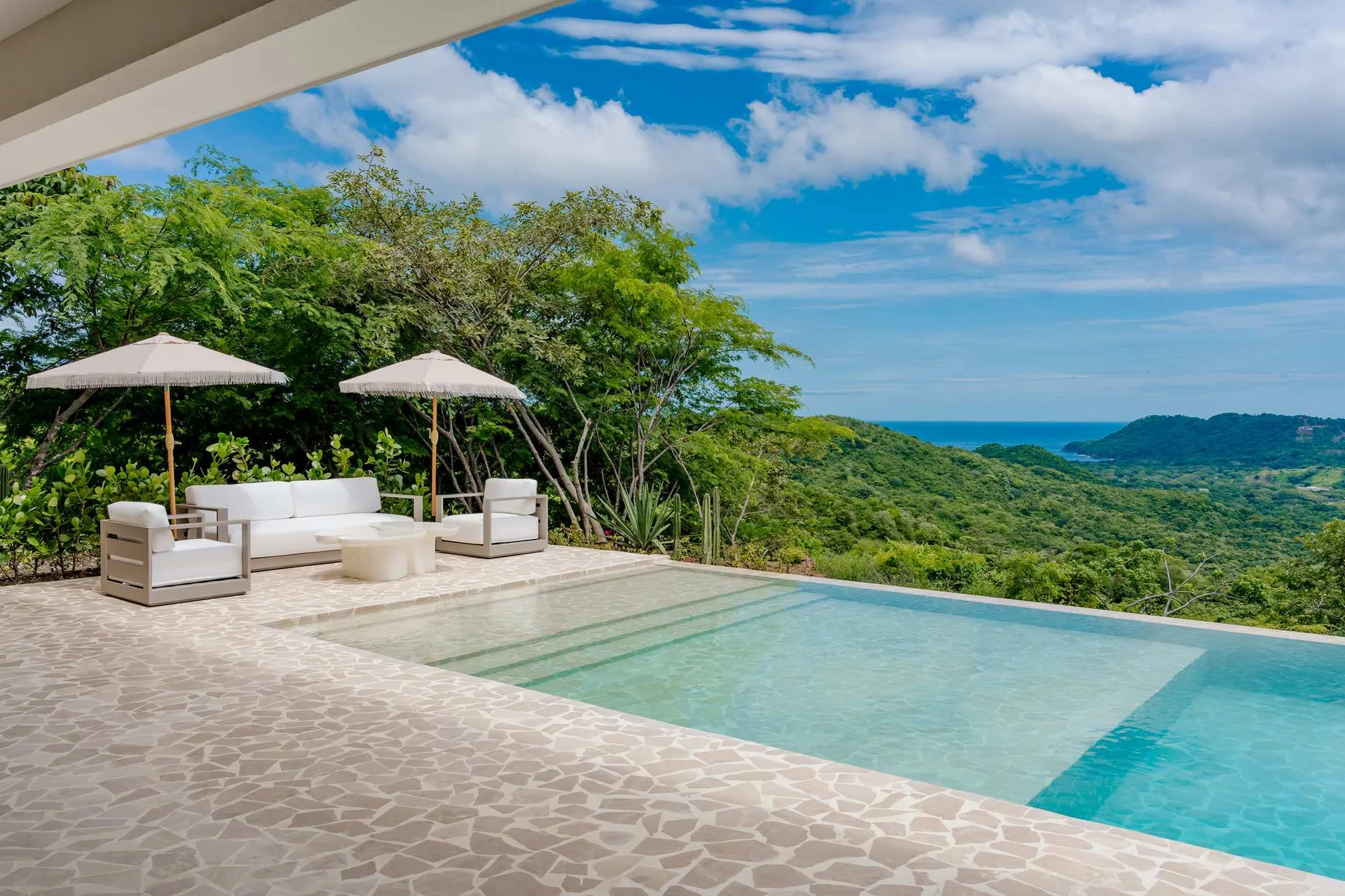 An outdoor swimming pool overlooks a green hillside and the ocean.