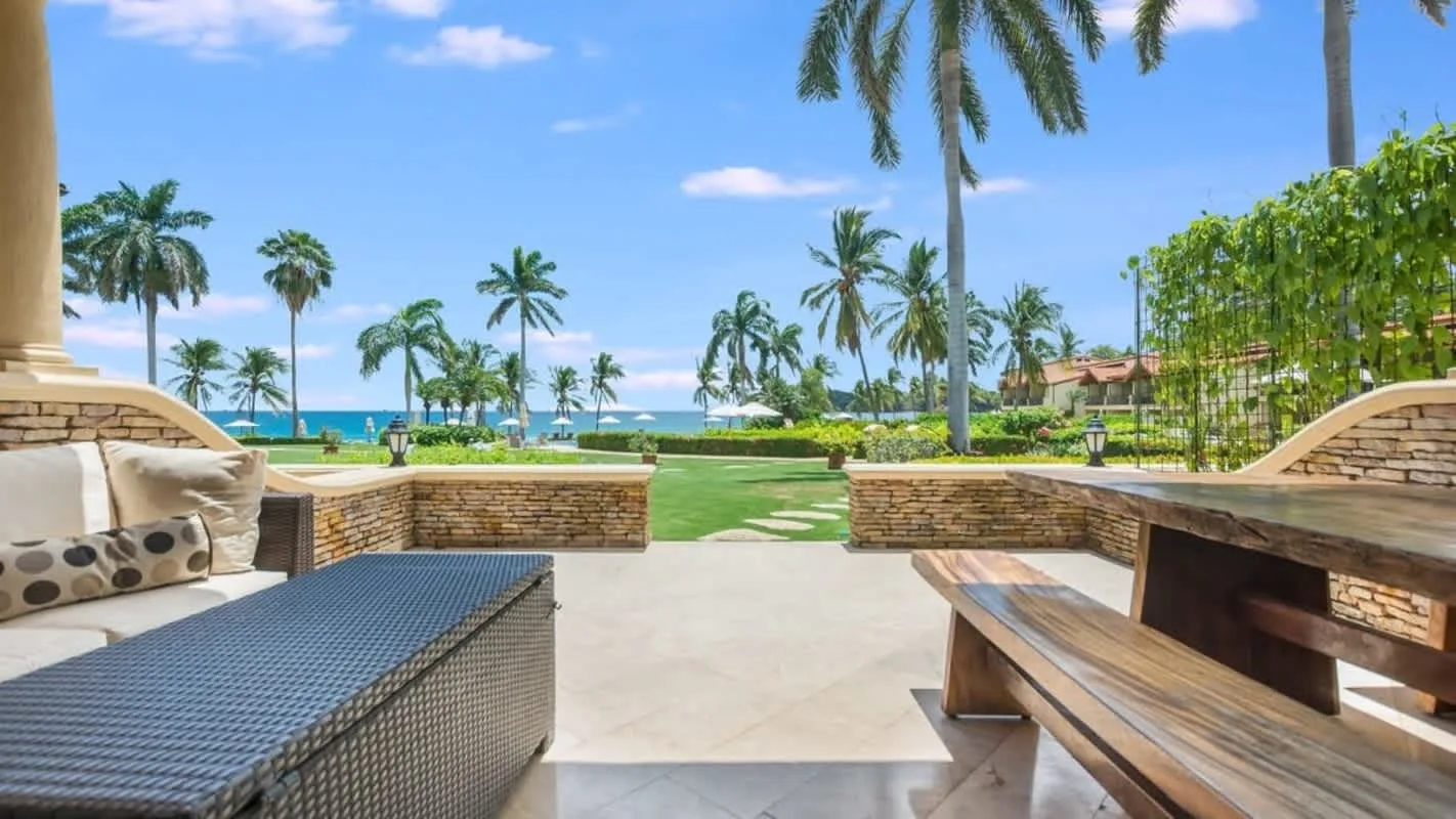 An outdoor patio with seating overlooks a landscaped lawn and palm-lined ocean view.