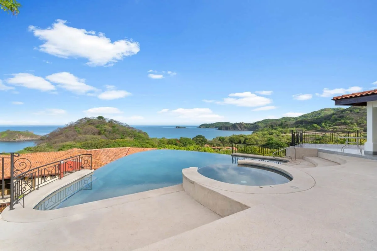 A rooftop pool overlooks green hills and the ocean under a bright blue sky.
