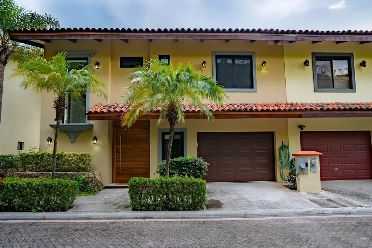 A yellow two-story house facade with palm trees and garage doors.
