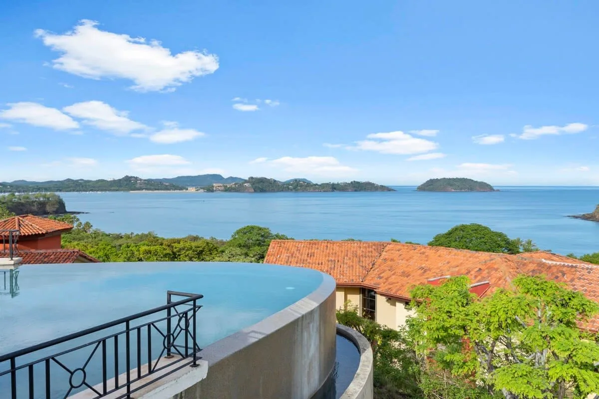 An outdoor pool overlooks the water, with tiled rooftops and green hills beyond.