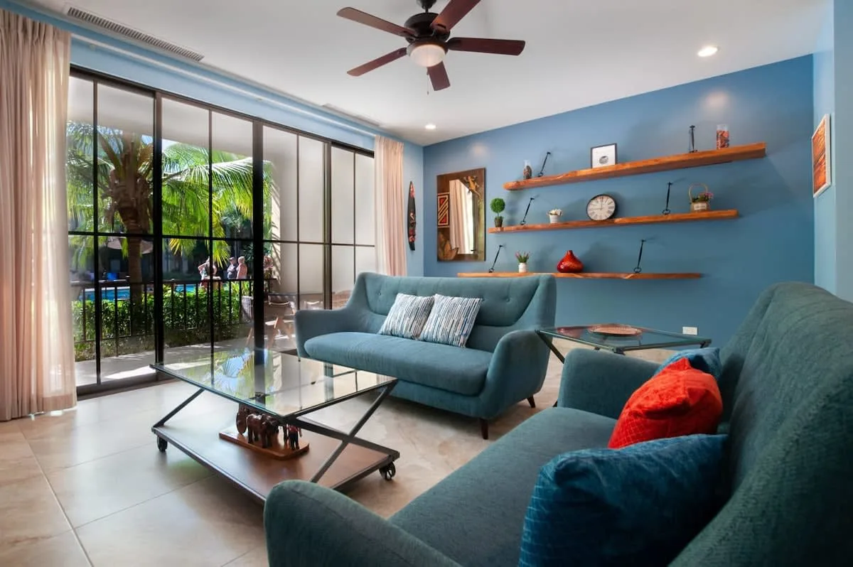 Una sala de estar con asientos azules, mesas de vidrio y puertas corredizas hacia la vegetación.