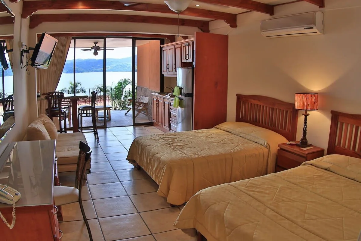 A bedroom with two beds, a bedside lamp, and an open view toward a balcony.