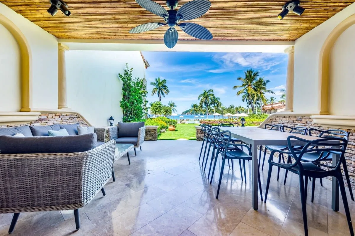 A covered outdoor dining and seating area opens onto a garden with palm trees.