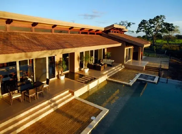 A modern building opens onto a pool, with a broad wooden deck and outdoor seating.