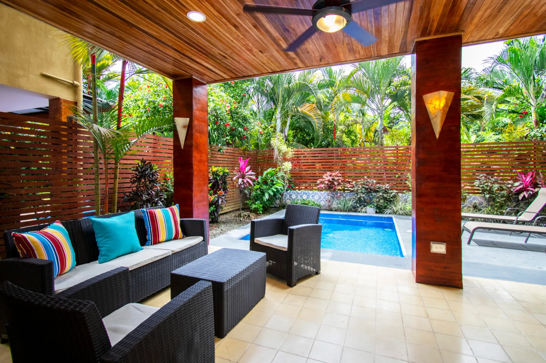 A covered patio has outdoor seating beside a small blue pool and tropical greenery.