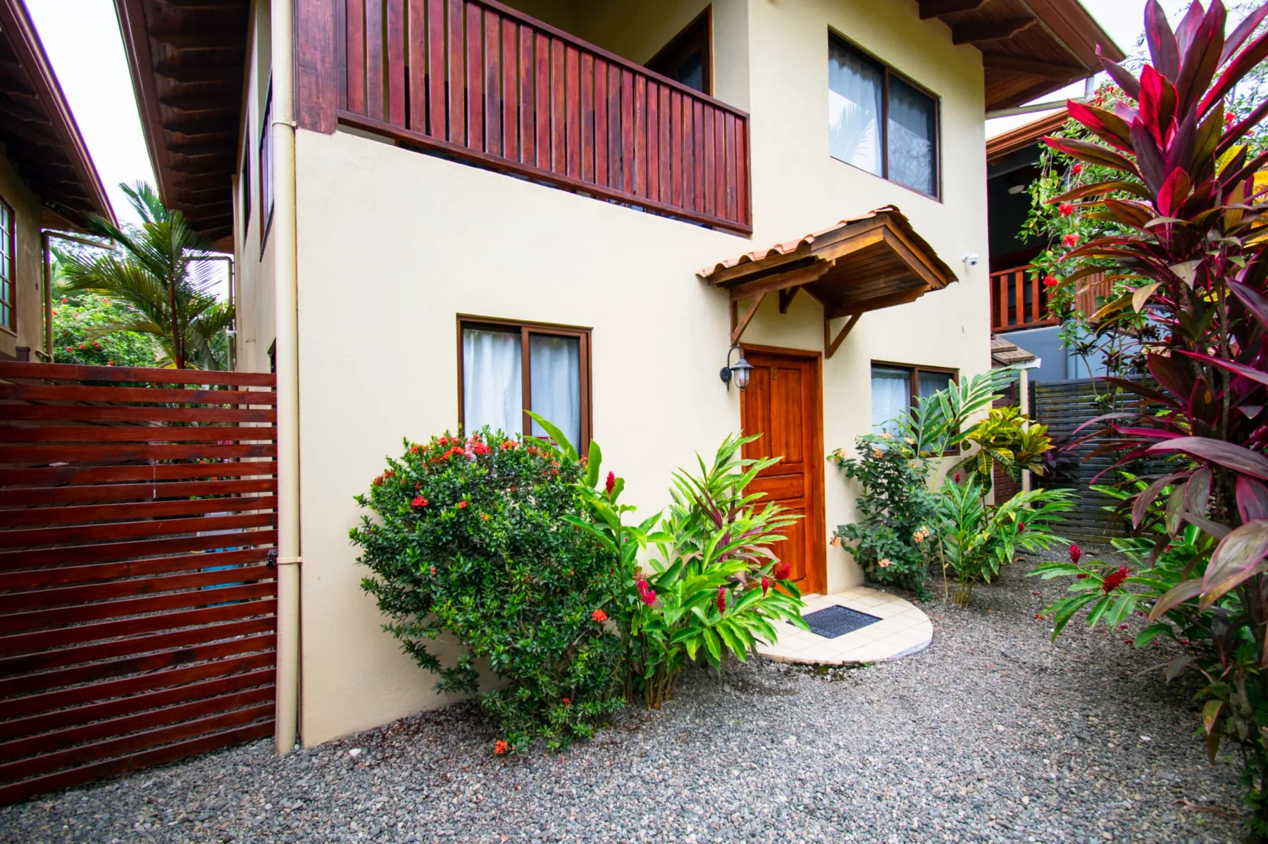 A house entrance opens onto a gravel courtyard surrounded by tropical plants.