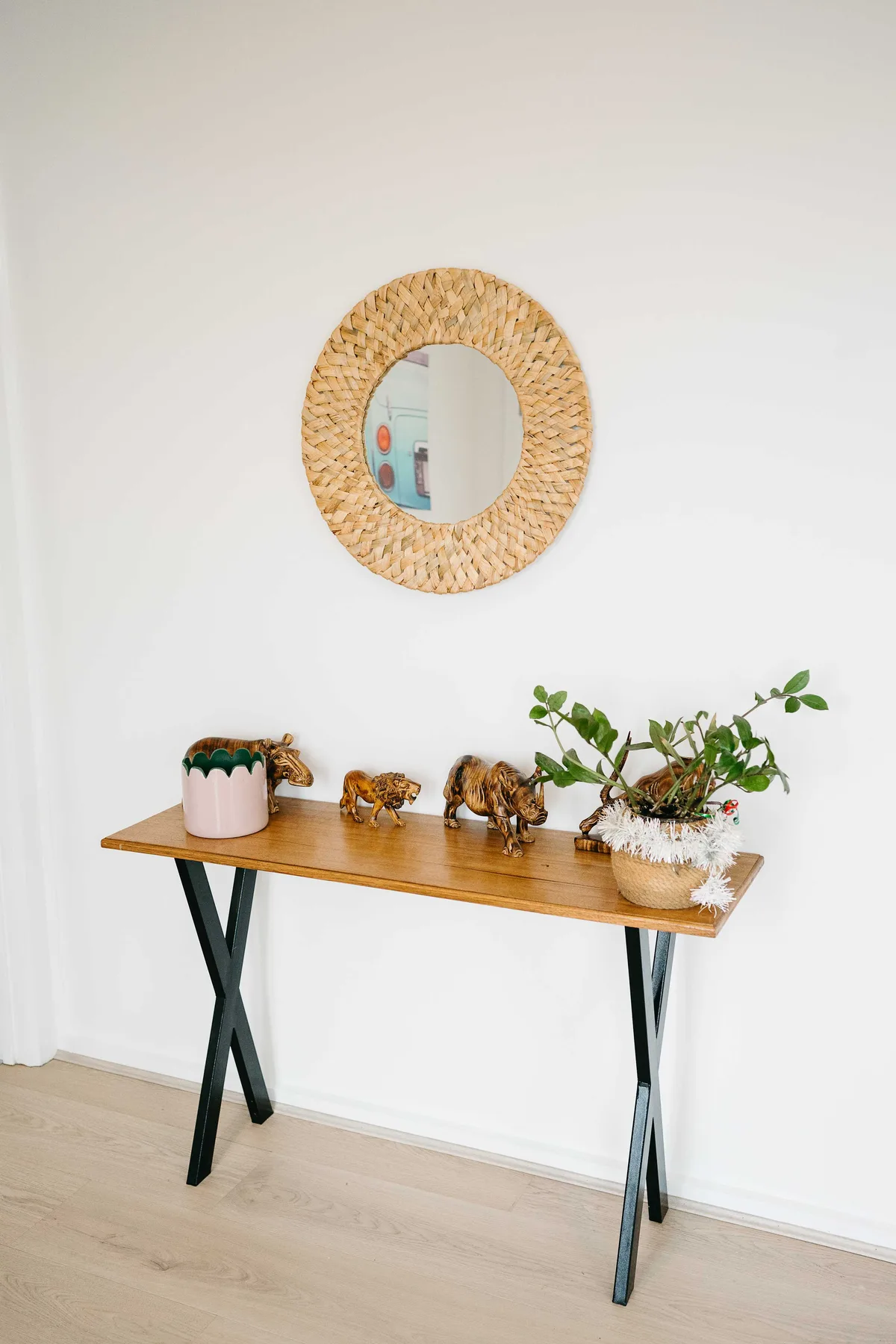 Una mesa consola estrecha con plantas y pequeñas figuras decorativas está junto a una pared blanca.