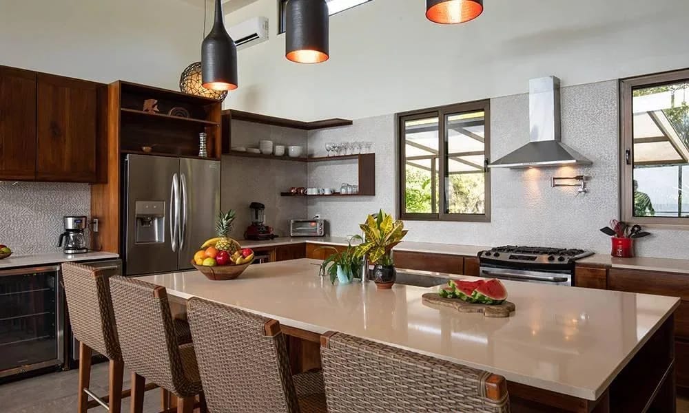 Casa Ola Vista · 11. This well-lit kitchen features dark wood cabinetry, a spacious central island with bar seating, stainless steel appliances, and large windows providing views of surrounding tropical foliage.