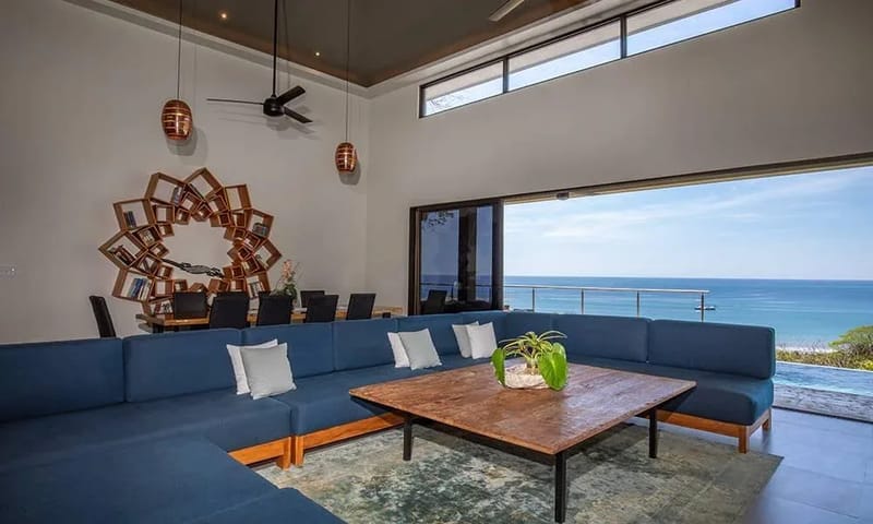 Casa Ola Vista · 13. An open-concept living space with high ceilings and a large sectional sofa features a dining area and wide glass doors that open to an expansive ocean view and an infinity pool.