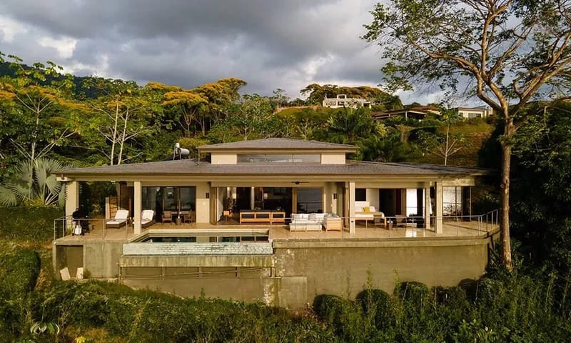 Casa Ola Vista · 9. The single-level villa features a broad covered terrace with a visible pool, outdoor lounging, and expansive glass doors opening to a lush tropical setting.