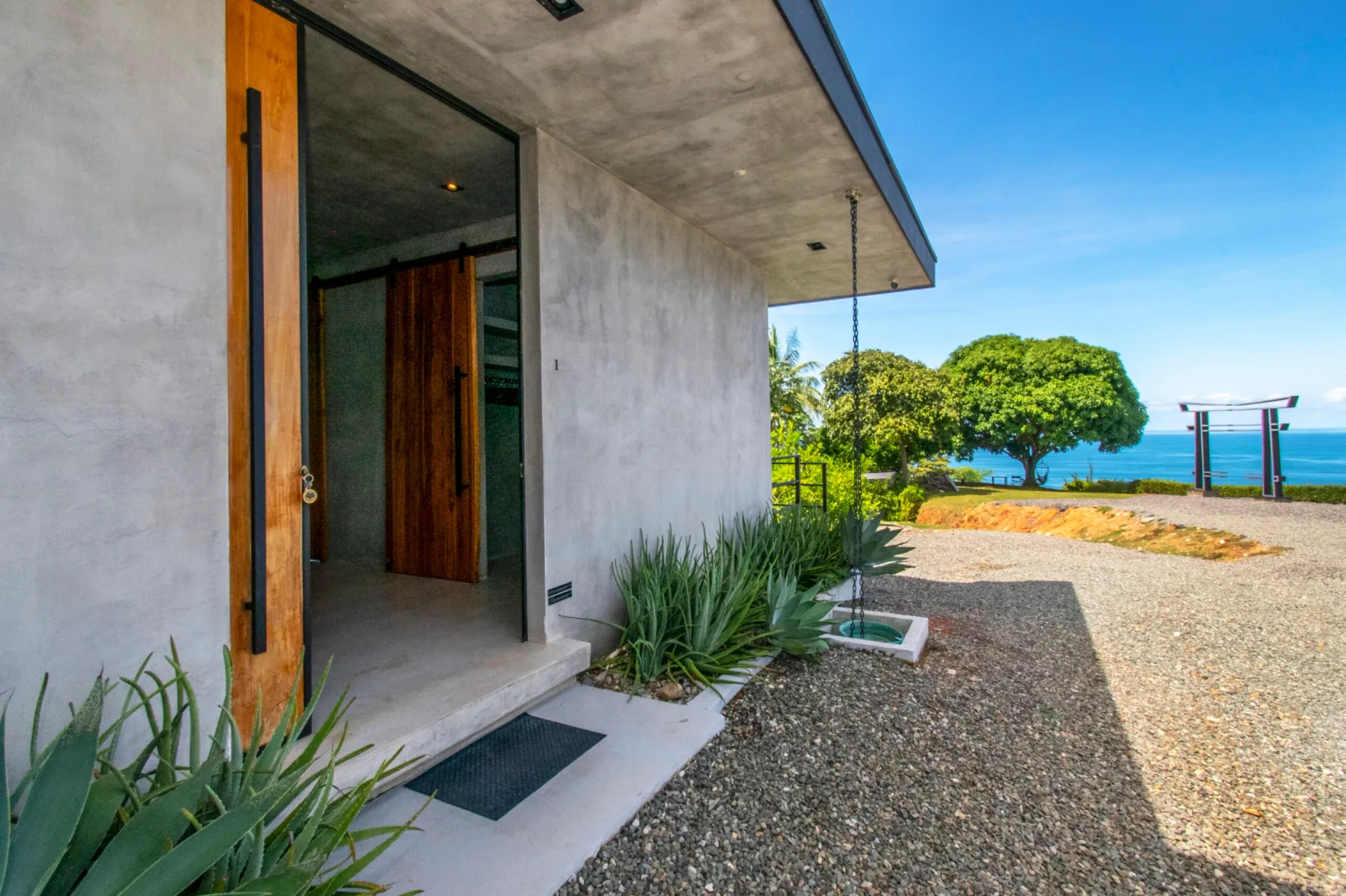 A concrete house entrance opens onto a gravel path with trees and water beyond.