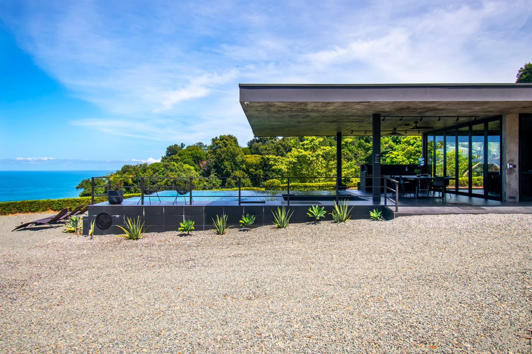 A modern glass-fronted building overlooks the ocean from a gravel terrace.