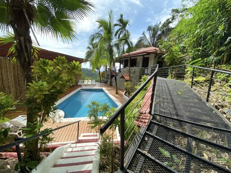 Casa Blue Wind · 17. The inviting blue swimming pool with a surrounding wooden deck and sun loungers is set amidst lush tropical foliage, offering views of green hills and access to the villa through multiple pathways.