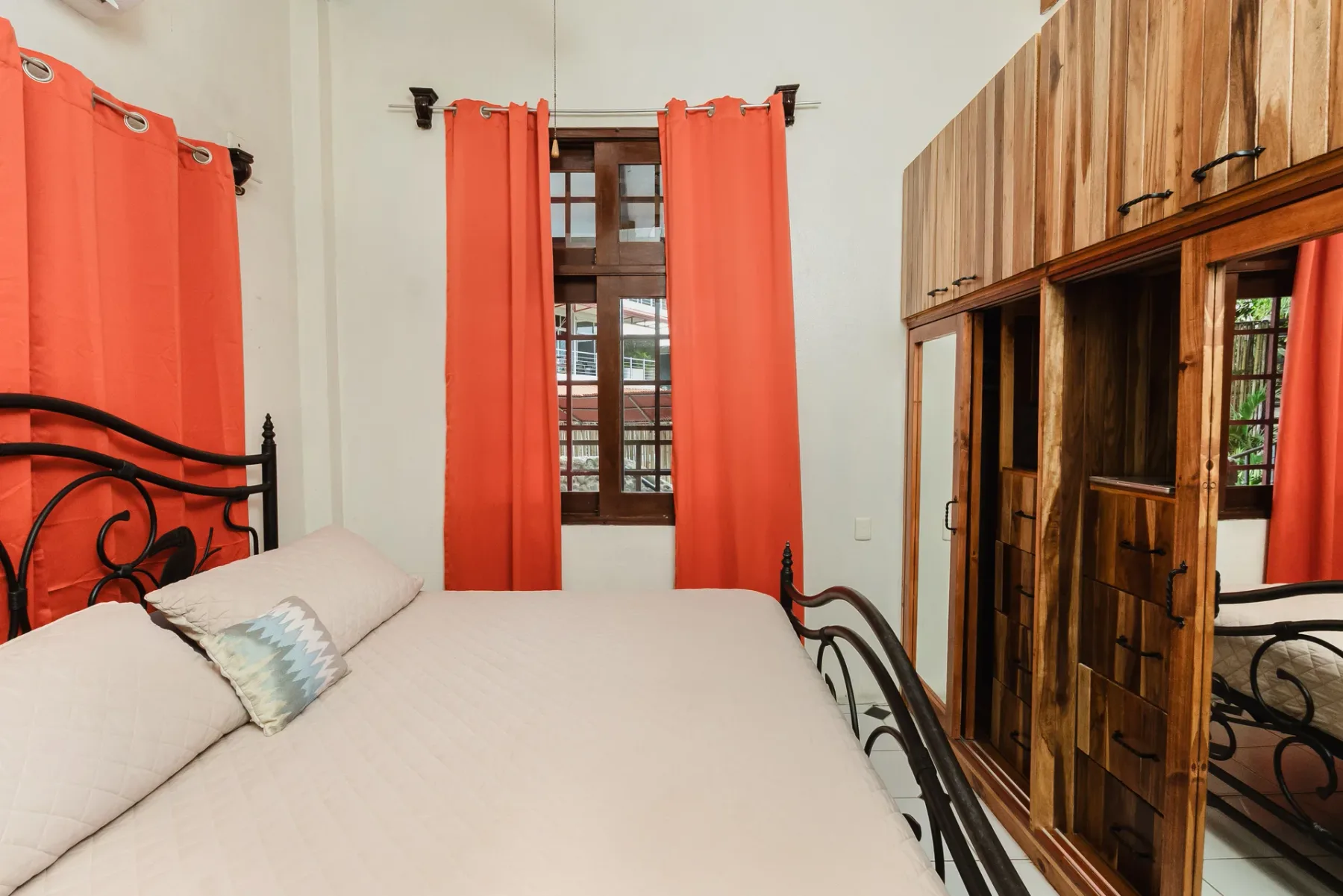 Casa Blue Wind · 14. A bright bedroom combines a wrought-iron bed, natural light from wood-framed windows draped with vibrant orange curtains, and rich wooden cabinetry.