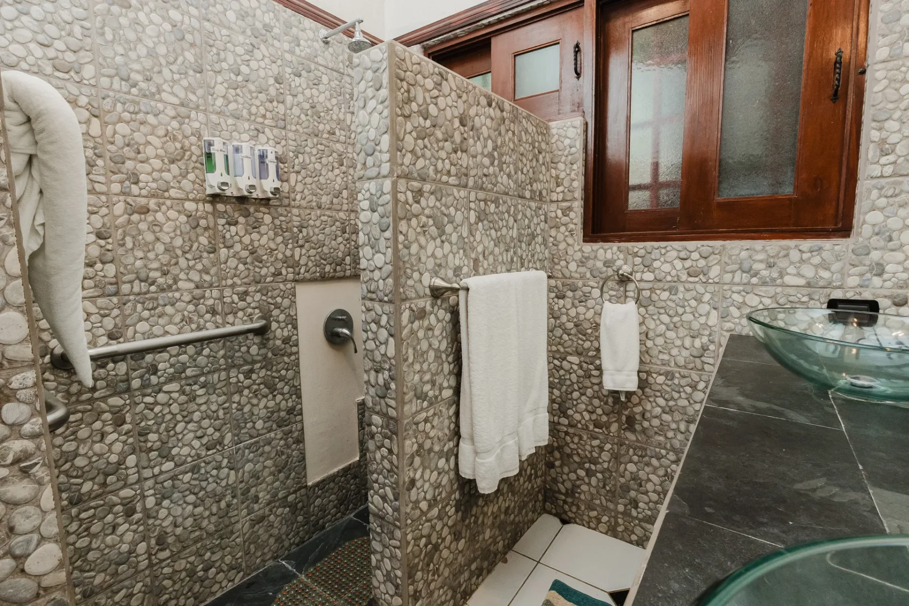 Casa Blue Wind · 19. This bathroom features natural pebble-tiled walls, a rain shower, a glass vessel sink, and a privacy window, creating a serene, earthy atmosphere.