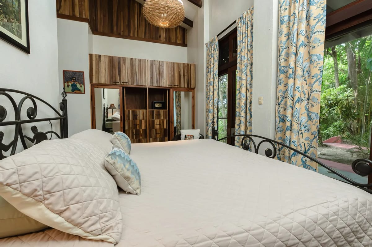 Casa Blue Wind · 2. This bright bedroom features a high beamed ceiling with a woven light fixture, a large metal-framed bed, built-in wooden storage, and a glass door looking out to tropical greenery.