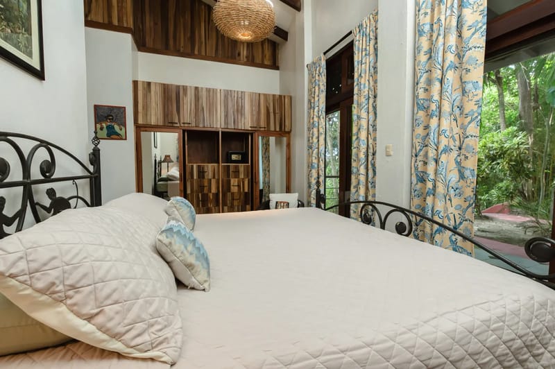 Casa Blue Wind · 2. This bright bedroom features a high beamed ceiling with a woven light fixture, a large metal-framed bed, built-in wooden storage, and a glass door looking out to tropical greenery.