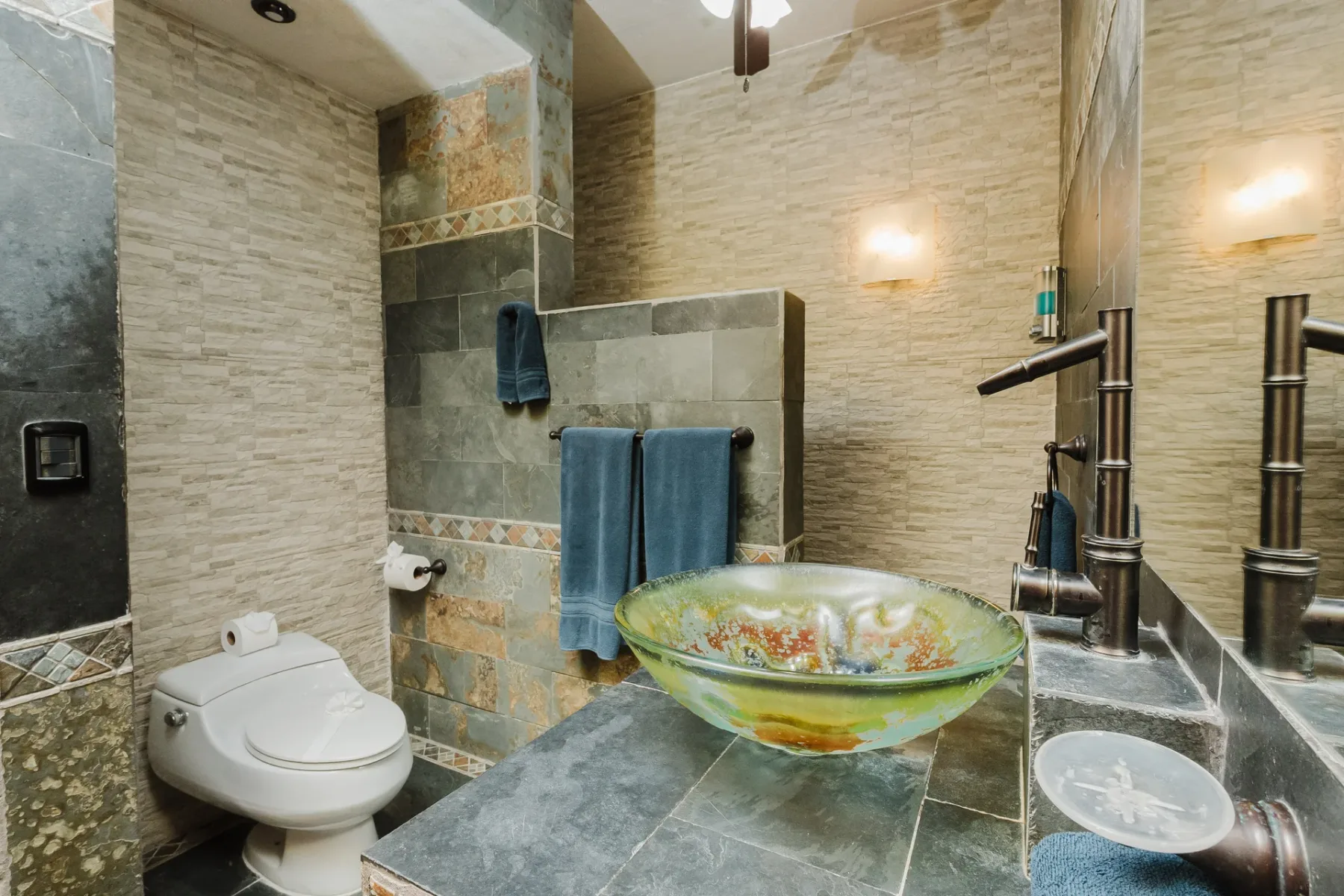 Casa Blue Wind · 15. A bathroom features textured stone and tile walls, a dark slate vanity with a distinctive glass vessel sink, a bamboo-style bronze faucet, and a toilet.