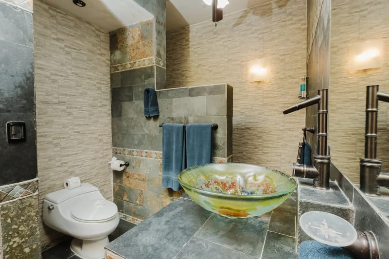 Casa Blue Wind · 15. A bathroom features textured stone and tile walls, a dark slate vanity with a distinctive glass vessel sink, a bamboo-style bronze faucet, and a toilet.