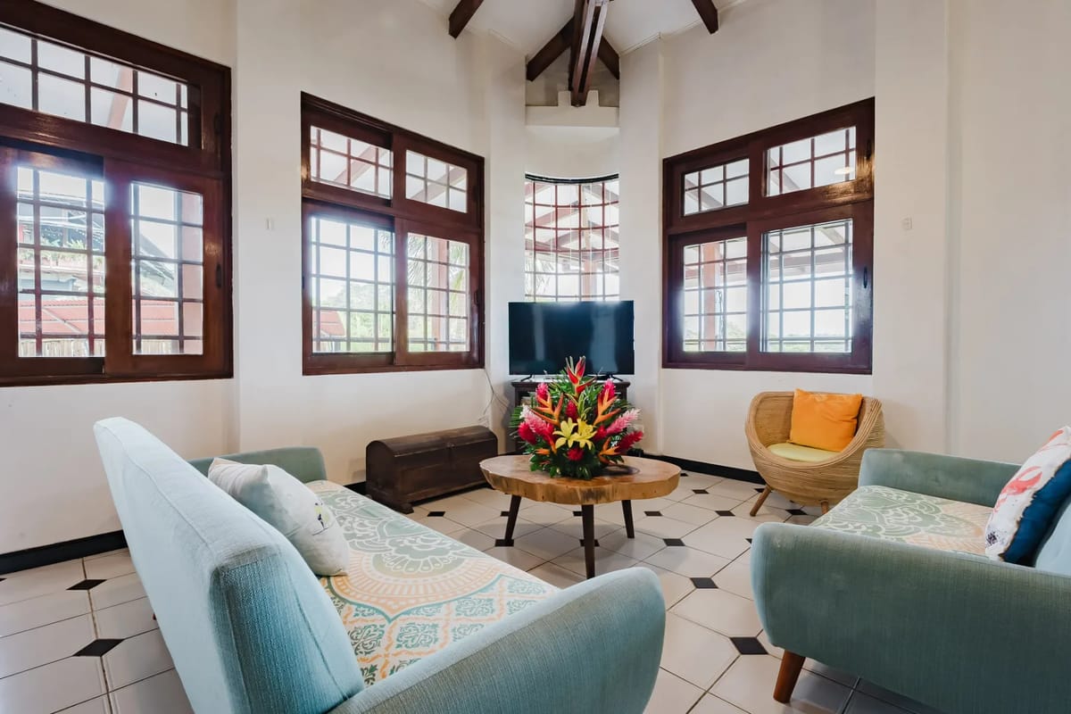 Casa Blue Wind · 4. A bright living room features a high vaulted ceiling with exposed wooden beams, multiple large grid-paned windows offering natural light and views of tropical greenery, and seating on light blue upholstered furniture.