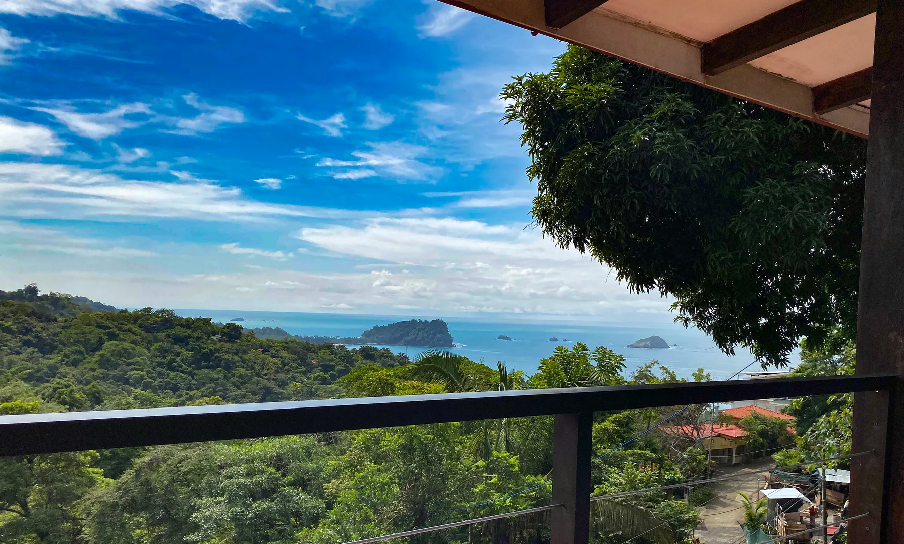 Casa Blue Wind · 8. From a shaded terrace, a wide view extends over lush tropical jungle to the Pacific Ocean, revealing several small islands and a bright, cloud-streaked sky.