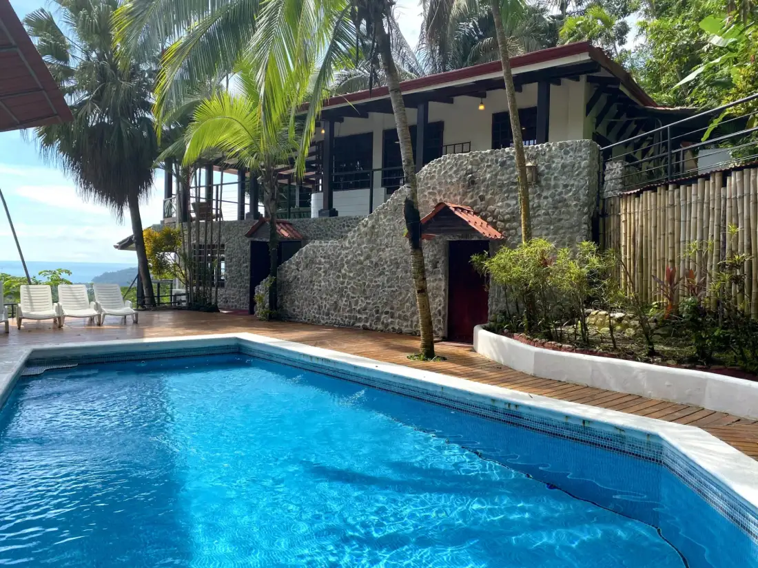 Casa Blue Wind · 28. An inviting blue swimming pool with a surrounding wooden deck and lounge chairs overlooks tropical greenery, the villa's stone and wood architecture, and a distant ocean view under bright daylight.