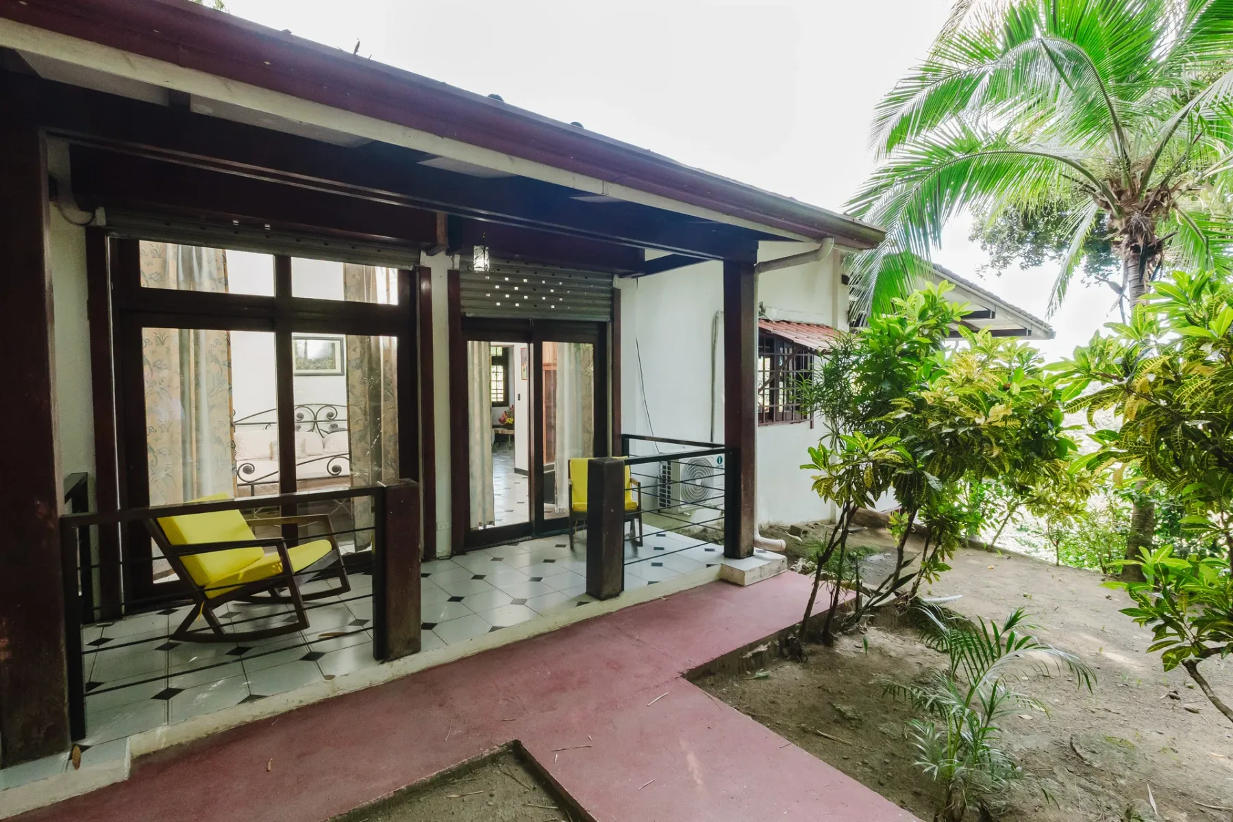 Casa Blue Wind · 26. A bright, covered outdoor terrace with dark wood columns and tiled flooring provides an inviting seating area, overlooking a lush tropical garden with a large palm tree.