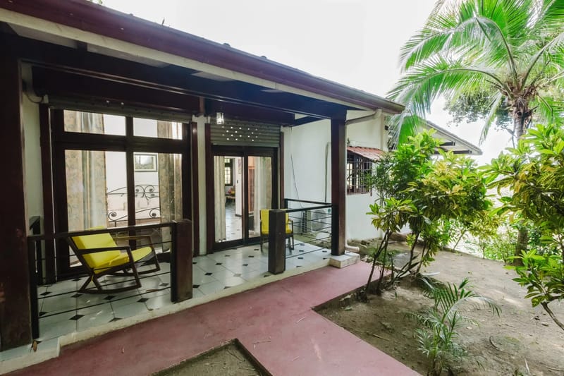 Casa Blue Wind · 26. A bright, covered outdoor terrace with dark wood columns and tiled flooring provides an inviting seating area, overlooking a lush tropical garden with a large palm tree.