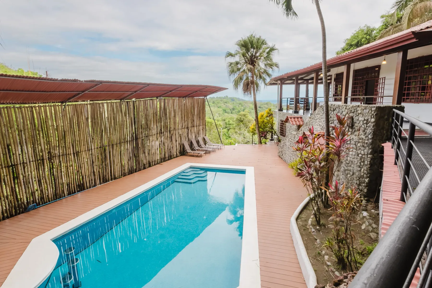 Casa Blue Wind · 27. A private swimming pool with steps is surrounded by a wood deck and tropical landscaping, featuring a villa with a stone facade and an upper-level terrace overlooking distant ocean and lush jungle.
