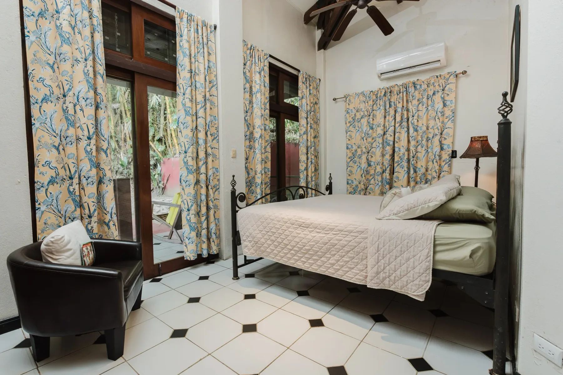 Casa Blue Wind · 25. An air-conditioned bedroom features a wrought iron bed, black and white tiled floor, exposed wooden ceiling beams, and large glass doors providing natural light and a view of tropical foliage.