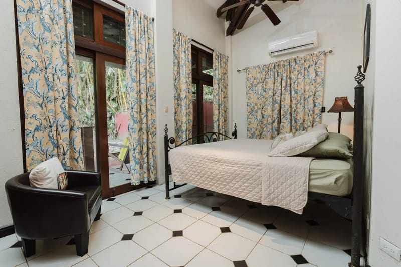 Casa Blue Wind · 25. An air-conditioned bedroom features a wrought iron bed, black and white tiled floor, exposed wooden ceiling beams, and large glass doors providing natural light and a view of tropical foliage.