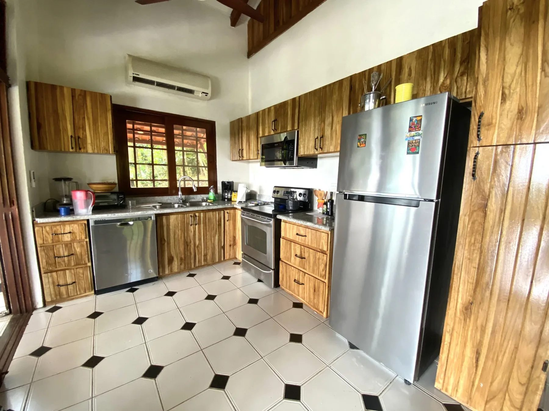 Casa Blue Wind · 16. This kitchen features rich natural wood cabinetry, a patterned tiled floor, stainless steel appliances, and a window above the sink offering views of tropical foliage and natural light, complemented by an overhead air conditioning unit.